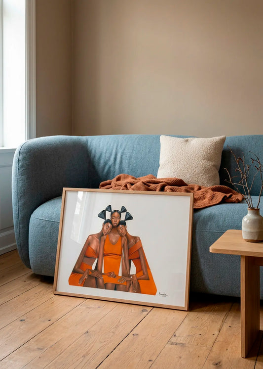 Orange Diamond Embrace by Manasseh Johnson Sr. – framed figurative art poster featuring three women in vivid orange clothing with black geometric diamond details, styled in front of a soft blue sofa in a warm neutral living room.