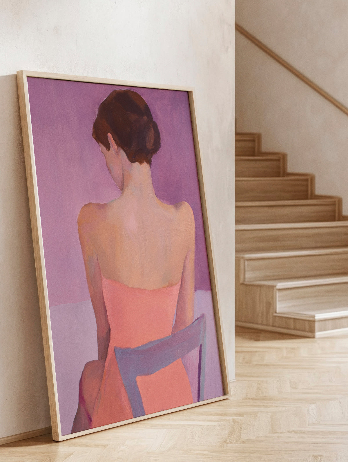 The Ballroom poster featuring a soft, painterly depiction of a woman seated with her back turned, in warm pink and lavender tones. Framed in light oak, the artwork leans elegantly against a textured wall near a wooden staircase, creating a serene and sophisticated ambiance.