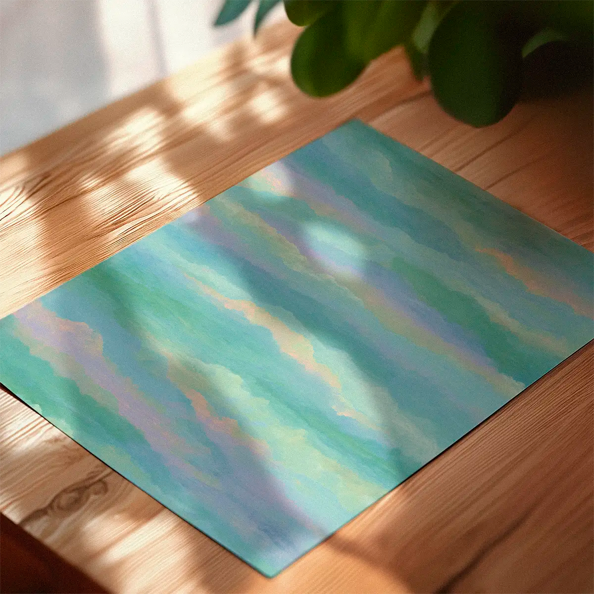 Soft pastel cloudscape poster lying on a sunlit wooden table, showing layered turquoise, lilac and peach tones.