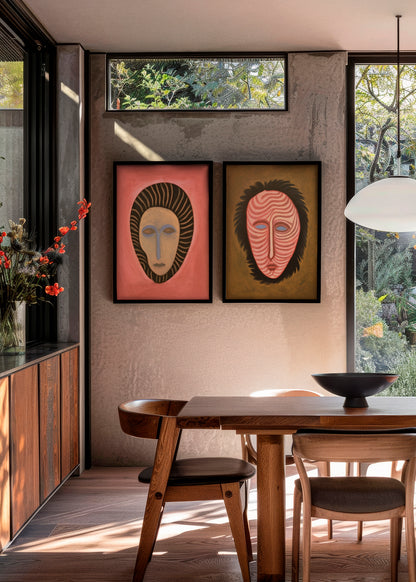 Framed posters Junglemen 1 and Junglemen 2 hanging side by side in a modern dining room with natural light, wooden furniture, and large windows. The left poster features a coral red background and striped mask motif, while the right one shows a mask with wavy lines on a golden brown background.