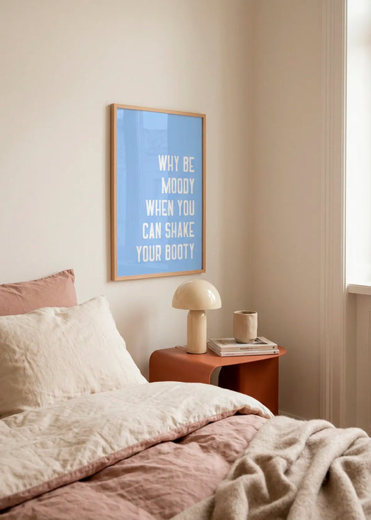 Shake Off the Blues by Athene Fritsch – framed blue typographic art poster with the words Why Be Moody When You Can Shake Your Booty, styled above a bedside table in a soft neutral bedroom.