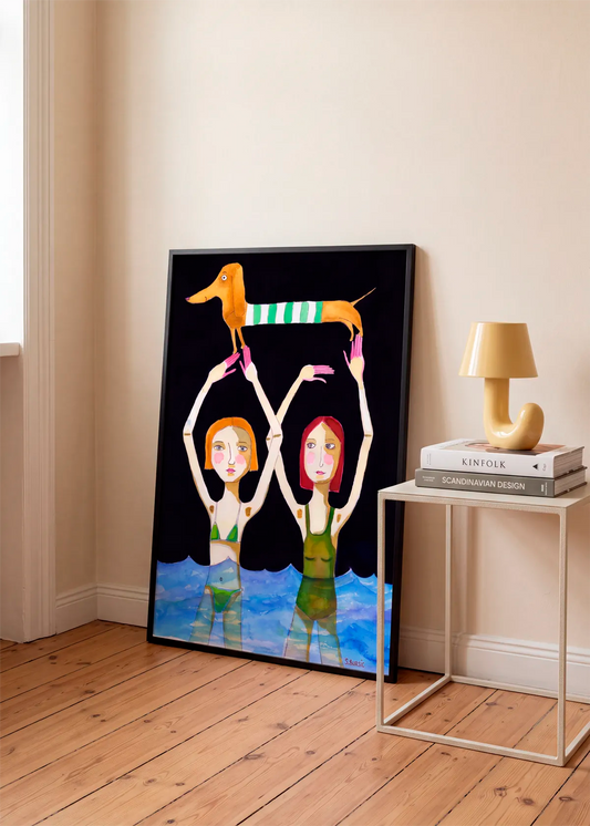 Whimsical Dog and Swimmers by Sharyn Bursic – framed art poster featuring two women in blue water holding a dachshund above their heads, styled in a warm neutral interior with wooden flooring.